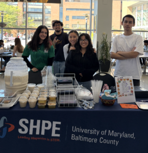 Group of SHPE UMBC students at a bake sale table with baked goods, drinks, and a menu displayed.